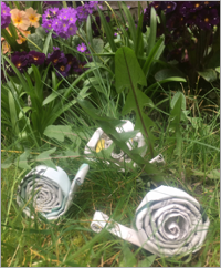 Three coiled up snails made of newspaper . They are among the plants in a garden.
