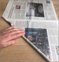 A hand rolling a newspaper into a tight tube on a wooden table.
